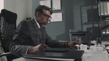 Masada çalışan kıdemli çift ırklı avukatın yan görüntüleri, dizüstü bilgisayardaki bilgileri kontrol etmek ve iş günlerinde notlar almak.