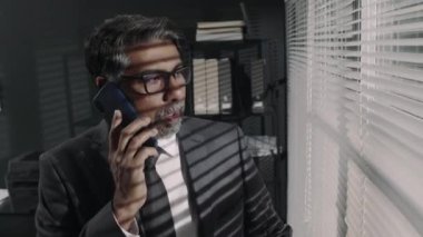 Üst düzey sakallı, takım elbiseli, gözlüklü bir avukatın müşteriler ve müşterilerle telefonda hukuki konuları tartışırken ve ofisteki panjurlardan gözetlerken görüntülerini göster.