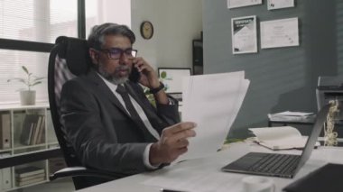 Takım elbiseli ve gözlüklü üst düzey erkek avukatın modern ofisteki müşteriyle akıllı telefondan konuşurken yasal belgeleri tartışırken çekilmiş yan görüntüleri.