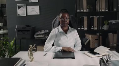 Ciddi bir Afrikalı Amerikalı kadın avukatın orta boy portresi. Gözlüklü ve ofis elbiseli. Modern ofisteki masada poz veriyor ve kameraya bakıyor.