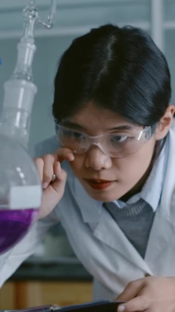 站在实验室里 看着紫色溶液的中国研究生的中等垂直率 检查反应或残留物 并在剪贴板上填写清单 — 图库视频影像