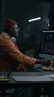 Bereli Afrikalı Amerikalı video editörünün dikey hali. Yapım sonrası şirket ofisinde tek başına oturuyor, bilgisayar ekranında yazılımda film görüntüleri üzerinde çalışıyor, kahve içiyor.