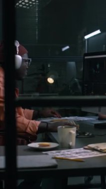 Siyahi erkek prodüksiyon editörünün orta dikey hali stüdyoda masasında oturuyor, profesyonel yazılımda ham görüntüler üzerinde çalışıyor, film senaryosuna danışıyor ve notlar alıyor.