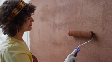Üzerinde sarı tişört ve kahverengi tulum olan gülümseyen beyaz bir kadının yan görüntüsü çocuk odasında boya silindiri kullanarak duvarı boyuyor.
