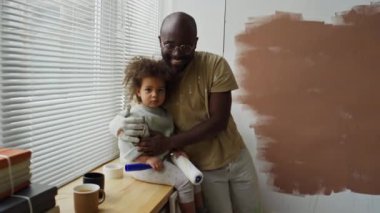 Minik melez kızına sarılan Afrikalı Amerikalı bir babanın portresi. Pencere kenarında oturmuş, tadilat sırasında kameraya bakarken gülümsüyor.