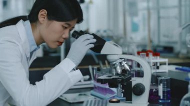 Beyaz önlüklü ve eldivenli genç Asyalı kadın bilim adamının mikroskop altında doku örneklerini eğip incelerken biyoteknoloji laboratuvarında tek başına çalışırken orta ölçekli bir fotoğrafı.