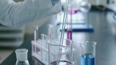 Tanımlanamayan laboratuar teknisyeninin uzun cam ampul pipetini pembe kimyasal reaktörle doldurup, temiz sıvıyı şişeye ekledikten sonra dairesel hareketle karıştırdığı eldivenli eldiven görüntüleri.