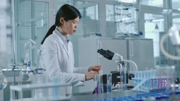 在实验室实验中 亚洲年轻女科学家走到工作台上的化学设备前 放下剪贴板 在显微镜下观察样品的中等镜头 — 图库视频影像