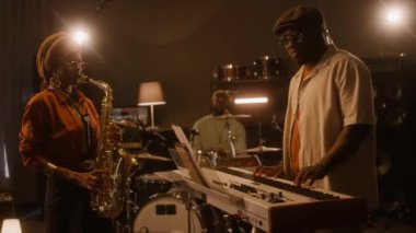 Orta boy, etnik türbanlı ve gözlüklü Afrikalı Amerikalı bir kadının saksafon çalarken, erkek grup arkadaşları klavye ve davul çalarken stüdyoda birlikte yeni müzikler yaparken.