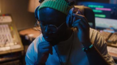 Bereli Afrikalı Amerikalı müzik yapımcısının orta ölçekli yakın çekimi, kulaklıkla müzik dinleyen gözlükler, tadını çıkarmak, ritme göre sallanmak, sonra ses ayarlarını ayarlamak için karıştırma masasına dönmek.