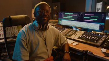 Bereli, gözlüklü, kulaklı neşeli Afrikalı Amerikalı müzik yapımcısının orta boy portresi dijital karıştırma masasının ve ekranın önünde oturuyor ve kayıt stüdyosunda yazılımla kameraya gülümsüyor.