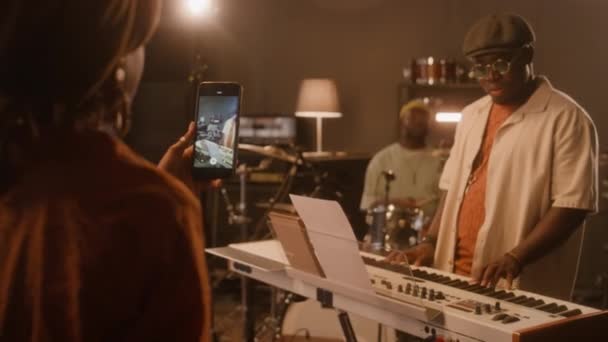 スタジオに立ってスマートフォンで撮影するターバンでアフリカ系アメリカ人女性バンド歌手のメディア 彼女の黒人の男性バンドメイトはキーボードやドラムを演奏しながら ギグのためにリハーサルしながら — ストック動画