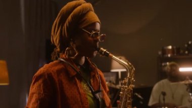 Etnik türbanlı ve gözlüklü Afrikalı Amerikalı bayan müzisyenin bandoyla prova yaparken ya da kulüpte konser verirken saksafon çalarkenki yakın çekimi.