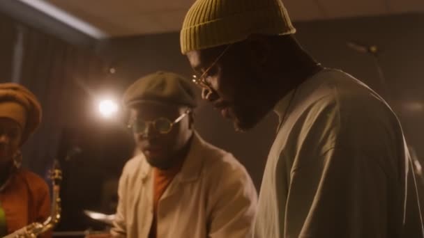 スタジオのキーボードに立っている2人のアフリカ系アメリカ人の男性バンドメイトのミディアムタイル張り 新しい曲のためのコードを試し アイデアを交換し サックスを見たり聞いたりする女性 — ストック動画