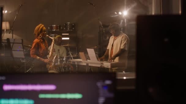 ハンドヘルドは レコーディングスタジオで演奏する黒人男性と女性のジャズミュージシャン およびオーディオ編集ソフトウェアを備えたコンピュータスクリーンを前景のサウンドエンジニアデスクで撮影した — ストック動画