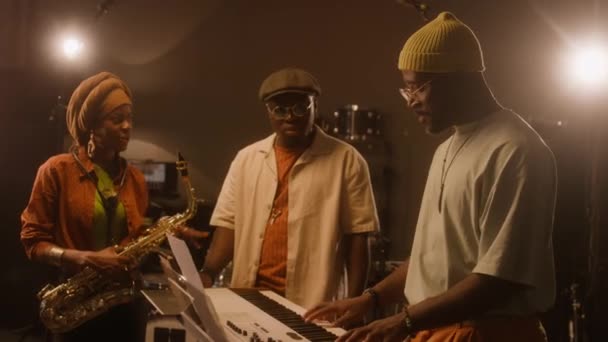 アフリカ系アメリカ人の男性と女性のブルースバンドのメンバーのメディアはスタジオのキーボードの周りに立っています メロディを構成する黒人男性 歌の気分で話すサックスフォンを持つ女性 — ストック動画