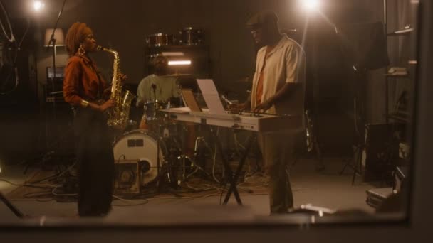 フルハンドヘルド コントロールルームウィンドウ アフリカ系アメリカ人のブルース男性と女性バンドのメンバーがサックス キーボード ドラムを演奏しながら レコーディングスタジオで一緒に演奏 — ストック動画