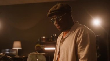 Genç Afrikalı Afrikalı Amerikalı müzisyenin şapkalı ve güneş gözlüklü halinin orta derece eğilişi, erkek grup arkadaşı davul çalıyor, yeni müzik kaydediyor veya stüdyoda birlikte prova yapıyor.