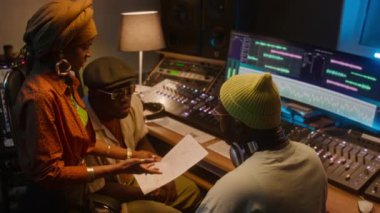 Türbanlı genç Afrikalı Amerikalı kadın şarkıcının orta sınıf yüksek açılı bir görüntüsü. Karıştırma tahtasında duruyor, grup arkadaşı ve yapımcısıyla şarkı sözlerini tartışıyor, stüdyoda yeni albüm kaydediyor.