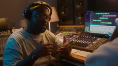 Orta omuzlu, kulaklıklı, kayıt stüdyosunda oturan, plak yapımcısıyla konuşan ve yeni şarkıları geliştirmenin yollarını tartışan neşeli Afrikalı Amerikalı erkek ses mühendisi.