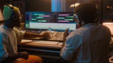 İki Afrikalı Amerikalı erkek müzisyenin stüdyoda birlikte çalıştığı, kulaklık taktığı, yeni parça dinlediği, yapımcı yönetmenlik mühendisinin frekans ve çıkış ayarlarını ayarladığı bir ortam.