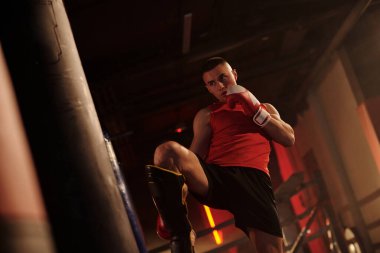 Genç profesyonel boksör antrenman sırasında, kum torbasının önünde dikilirken ve koruyucu önlem olarak bacağından vururken düşük tekme pratiği yapıyor.