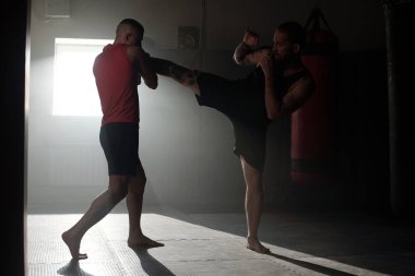 Kırmızı yelekli genç boksör, düşük tekme antrenmanı sırasında önünde duran siyah spor kıyafetli rakibini yakalıyor.