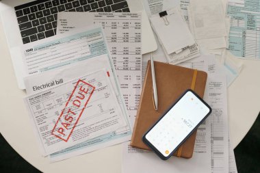 Dizüstü bilgisayarın klavyesinin genel görünümü, süresi geçmiş elektrik faturası, kahverengi deri kapaklı defter ve ekranında hesap makinesi olan akıllı telefon.