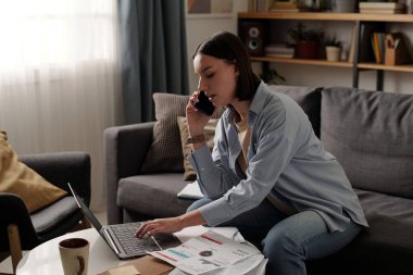 Oturma odasındaki kanepede oturan, dizüstü bilgisayarın tuşlarına basan ve cep telefonuyla konuşan gündelik kıyafetli genç bir kadın.
