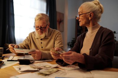 Beyaz sakallı ve saçlı yaşlı bir adam karısının yanında oturmuş banknotları sayarken mali faturaları inceliyor.