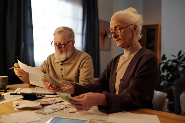 さまざまな住宅支払い手数料と彼女の夫に対するドルの紙幣を数えるテーブルで座っている眼鏡の深刻な年齢の女性 ストック画像