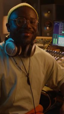 Bereli, gözlüklü, kulaklıklı gülümseyen Afrikalı Amerikalı müzik mühendisinin dikey portresi kayıt stüdyosunda miksaj ve düzenleme ekipmanlarının önünde oturmuş kameraya bakıyor.