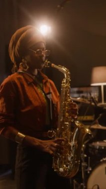 Etnik türbanlı ve gözlüklü Afrikalı Amerikalı kadın caz grubu üyelerinin konserde veya stüdyoda prova yaparken saksafon çalarken orta dikey görüntüsü.