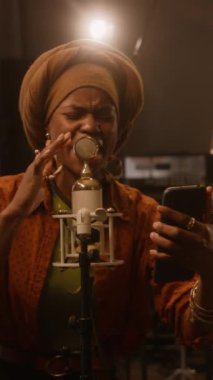 Renkli etnik türbanlı Afrikalı kadın caz şarkıcısının orta dikey görüntüsü stüdyoda retro mikrofonda şarkı söylemesi, akıllı telefonda çekim yapması veya yayını yapması, bandoyla prova yapması.