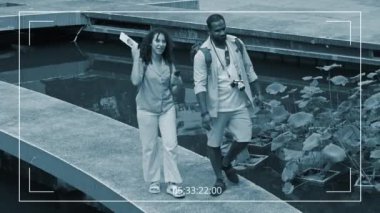 Güvenlik kamerası, Afrika kökenli Amerikalı bir adam ve beyaz bir kadının sırt çantalı, turist broşürleri ile birlikte gölette yürürken, gülümserken, sohbet ederken, yurt dışında tatilin tadını çıkarırken çekilmiş.