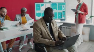 Hippi kıyafetli, sarı spor ayakkabılı, tekerlekli sandalyede oturan dijital pazarlama şirketinin yaratıcı atmosferinde dizüstü bilgisayarda hızla yazan genç Afrikalı Amerikalı adamın tam görüntüsü.