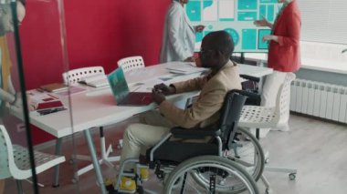 Çeşitli yaratıcı çalışanların toplantıda proje tartışması çekimleri, tekerlekli sandalyedeki siyah adam ve çift ırklı meslektaşın dizüstü bilgisayar, tablet, ekrandaki baskıya bakan kadınlar.