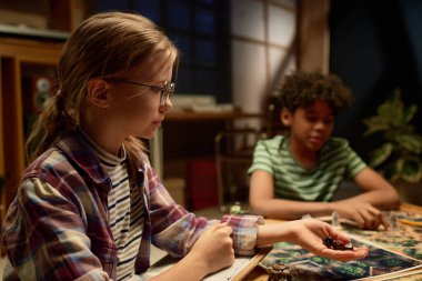 Gözlüklü genç bir kızın oyun tahtasına bakarken arkadaşlarının yanında oturup zar atmasının yan görüntüsü.