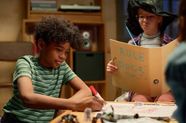 Çizgili tişörtlü yakışıklı Afro-Amerikan çocuk masanın yanında oturuyor ve kırmızı fosforlu kalemle oyunun kurallarının altını çiziyor.