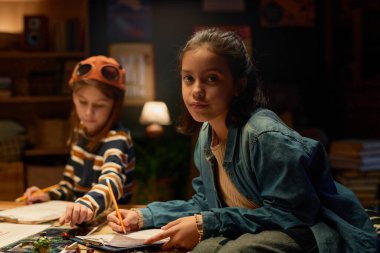 Mavi kot gömlekli ya da ceketli şirin Latin kız kameraya bakıyor ve arkadaşına karşı kalemle masa üstü oyununun kurallarını işliyor.