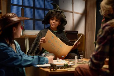 Cadı şapkalı genç Afro-Amerikan çocuk elinde açık oyun kuralları kitabını tutuyor ve önünde oturan arkadaşlarına anlatıyor.