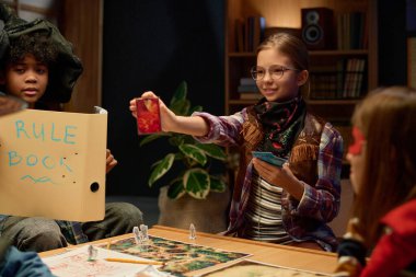 Oyun tahtası ve karakter figürleriyle masada dururken arkadaşlarına kartlarından birini gösteren gözlüklü sevimli bir kız.