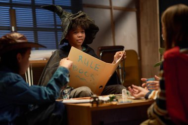 Cadı şapkalı ve siyah mantolu Afrikalı genç bir çocuk oyun tahtasıyla masada oturuyor ve bir grup arkadaşa kuralları anlatıyor.