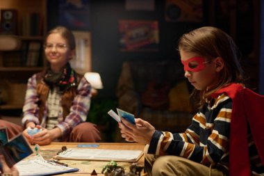 Kırmızı maskeli ve Süpermen 'in peleriniyle masanın başında oturmuş kızlara karşı masa başında oturan sevimli genç bir çocuk.