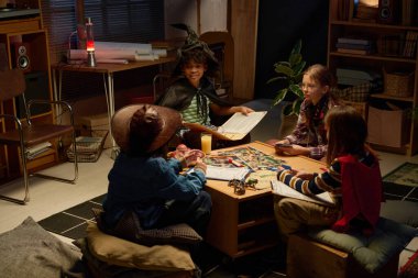 Dört kültürler arası arkadaş canlısı çocuk masa oyunu oynarken cadı mantolu ve şapkalı çocuk kuralları anlatıyor.