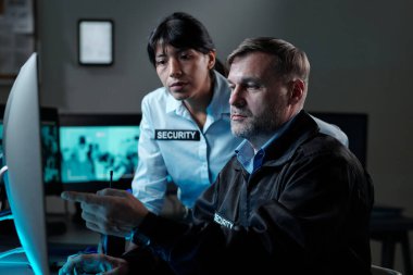 Üniformalı, kendine güvenen erkek gardiyan genç bayan meslektaşla video görüntülerini tartışırken bilgisayar ekranını işaret ediyor.