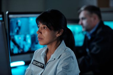 Güvenlik görevlisi üniformalı esmer bir kadın gözetleme odasında bilgisayar başında oturup ekrana bakıyor.
