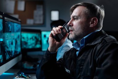 Ağızdan telsizle konuşan yetişkin bir erkek güvenlik görevlisi bilgisayar ekranının önünde otururken meslektaşına durumu bildiriyor.