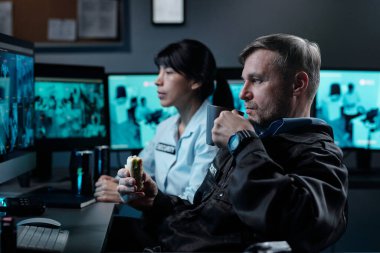 Ciddi bir yetişkin polis memuru bilgisayar ekranının önünde oturup kapalı devre televizyon izlerken bir fincan kahve ve sandviç yiyor.