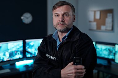Elinde telsizle gözetleme odasında bilgisayar monitörleriyle kameraya bakan ciddi bir yetişkin güvenlik görevlisi.